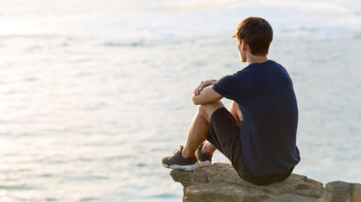 young man diagnosed with schizoid personality sitting alone on top of a cliff above the ocean view at sunset